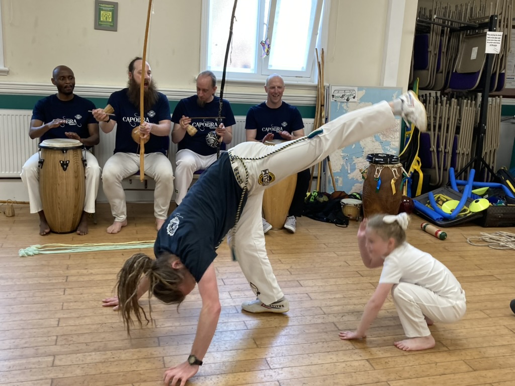 A man on the ground kicking over the head of a small girl. A group of people watch, playing instruments.