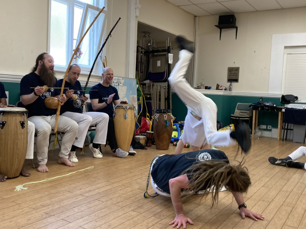 A man and child play capoeira while a group of musicians play and watch, laughing.